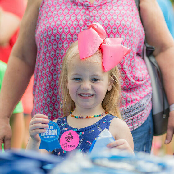 A little girl with a big, pink bow in her hair holding a blue ribbon and smiling at the camera during Ice Cream Days in Le Mars.